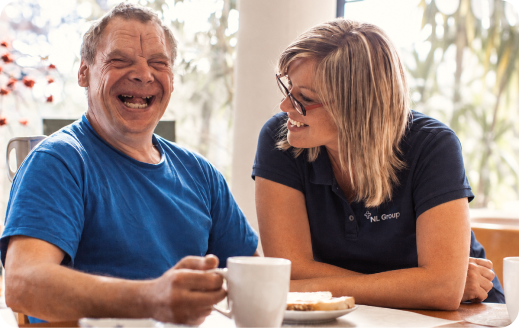 A man and a woman sit at a table with mugs and a plate of food, smiling and laughing together in a bright indoor setting.