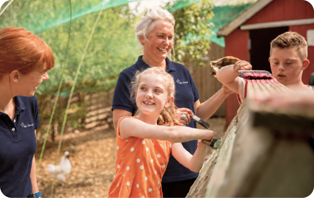 Three adults and a child clean a wooden surface outdoors on a sunny day, with a red shed and some trees in the background.