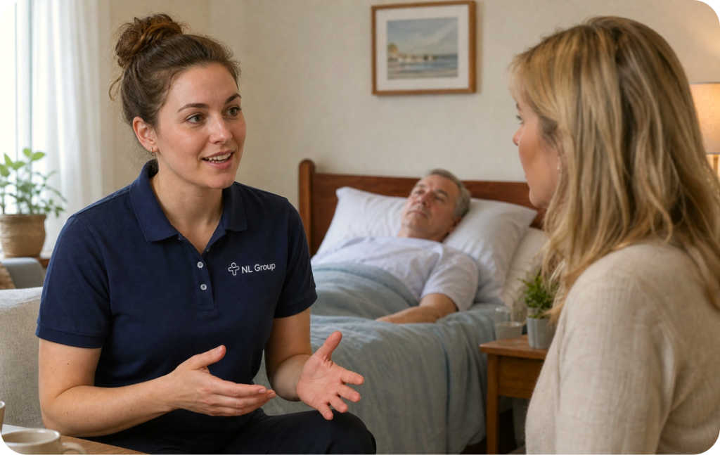 A woman in a navy polo shirt talks to another woman by a bedside, with an older man lying in the bed behind them in a home setting.