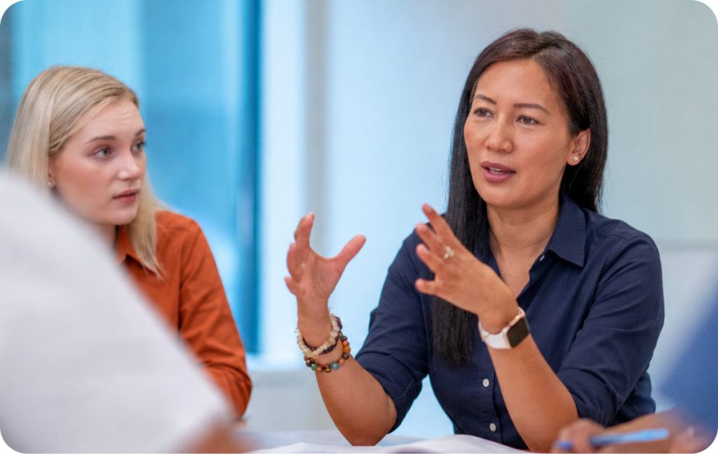 Two women sit at a table in discussion. One woman gestures with her hands while speaking, and the other listens attentively.