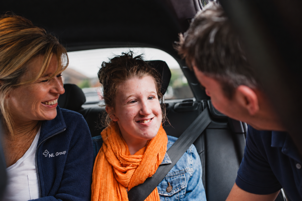 Three people sit in a car, two adults smiling at a young woman in the middle who is wearing a seatbelt and an orange scarf.