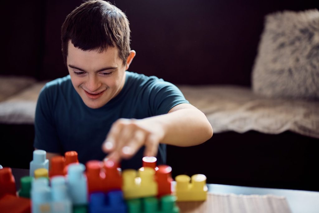 A young person smiles while assembling colorful plastic building blocks on a table in a cozy indoor setting.